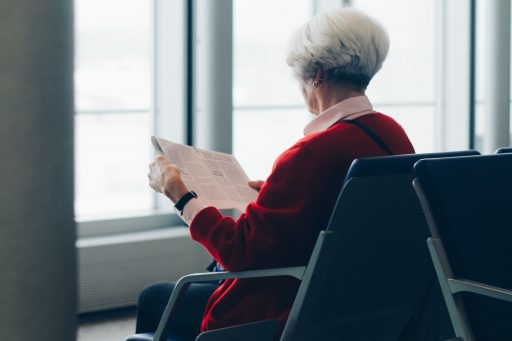 Grey haired woman sitting, waiting, and reading newspaper at airport waiting area.