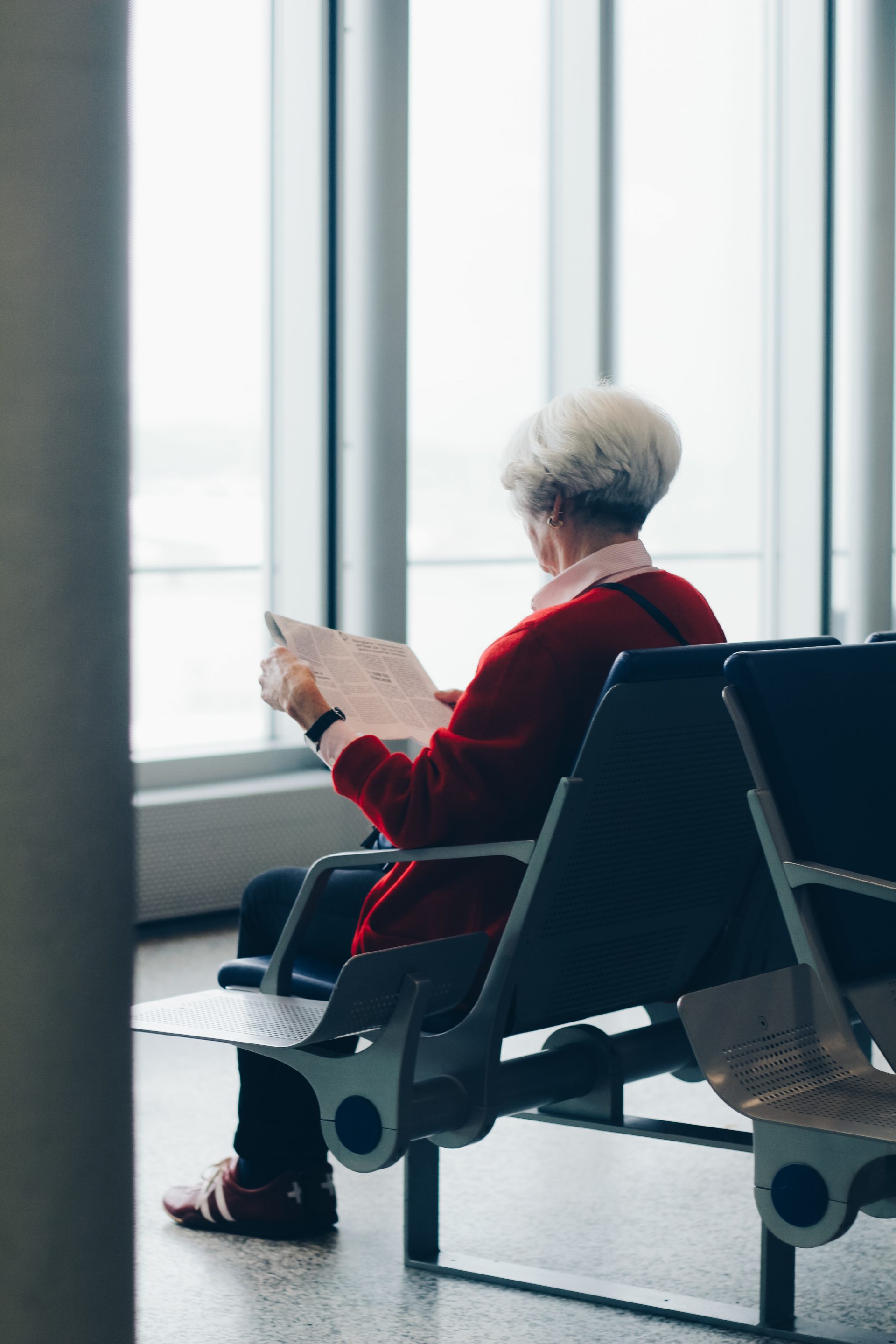 Grey haired woman sitting, waiting, and reading newspaper at airport waiting area.