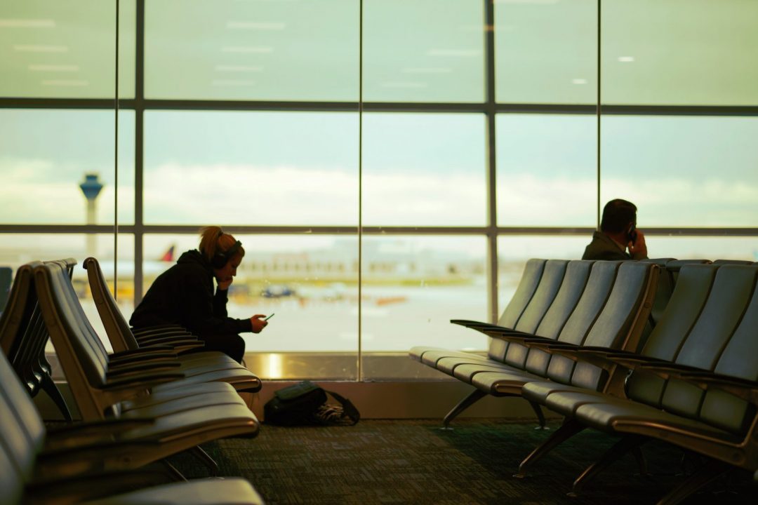 Sitting silhouettes against airport window, once checking phone for delayed flight info